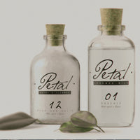 An advert for a fictional brand called Petal, showing Two old, clear, apothecary style glass bottles with cork lids, containing an essential oil
