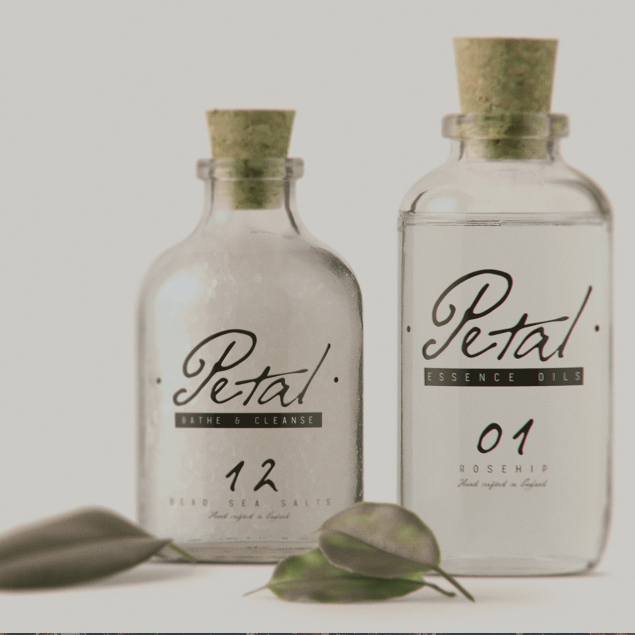 An advert for a fictional brand called Petal, showing Two old, clear, apothecary style glass bottles with cork lids, containing an essential oil